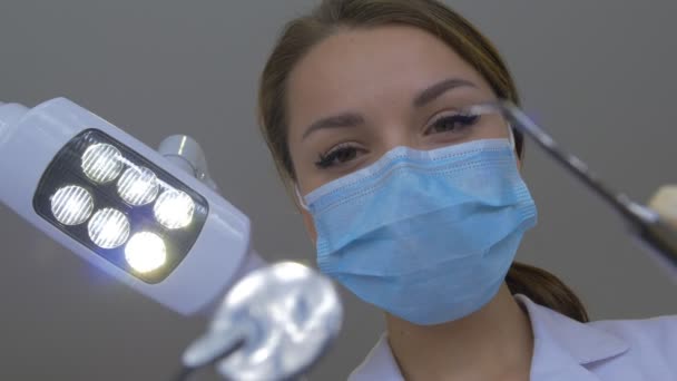 Jovem dentista em abordagens de máscara com ferramentas em pé sobre um paciente acende a luz em seguida, fora das mãos de revestimento de laboratório no exame de luvas na clínica dentária — Vídeo de Stock