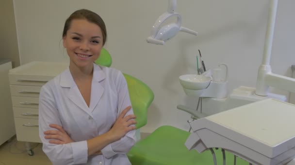 Mujer joven dentista en abrigo de laboratorio está sonriendo sentada con los brazos cruzados en una silla Máquina de dentista Lámpara dental Silla verde Paredes blancas Clínica dental — Vídeos de Stock