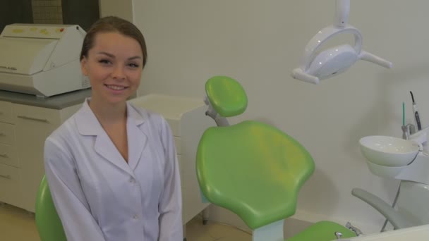 Jovem dentista convida para uma cadeira com mulher de mão em casaco de laboratório está sorrindo sentado na máquina de uma cadeira Dentista Lâmpada Dental Cadeira Verde Clínica Dental — Vídeo de Stock