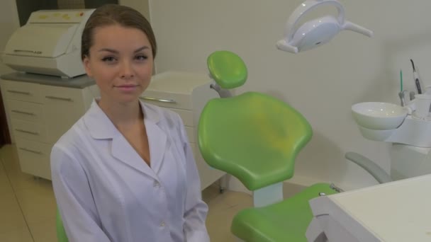 Joven dentista sonriente invita a la silla Mujer con abrigo de laboratorio está sentado en la máquina de un dentista de silla Lámpara dental Silla verde Clínica dental Sala — Vídeos de Stock