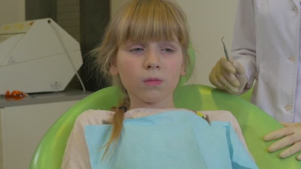 Kid is licking Her Lips Sitting at Dentist Chair Paciente sentado em uma cadeira verde Doctor is Holding a Suction and Hook Dental Clinic Visita a um dentista — Vídeo de Stock