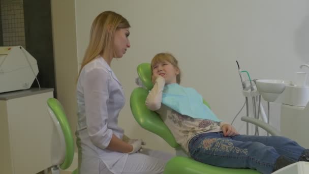 Zahnärztin spricht mit Kind, das die Wange berührt, und untersucht die Zähne eines Patienten. Kind sitzt in einem grünen Stuhl Zahnklinik-Besuch beim Stomatologen — Stockvideo