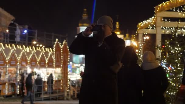 Hombre está tomando fotos Feria de Navidad Plaza de Sofía Kiev Noche de Año Nuevo en una plaza Mikhailovskaya Catedral de Sofía Cupolas de Oro Luces Kioscos Láseres — Vídeo de stock