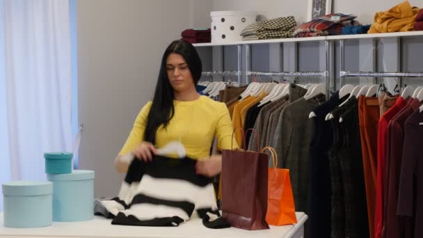 Mujer pliega una blusa Cliente vendedor está sonriendo Mujer ropa Tienda Morena Mujer está comprando una ropa en una boutique Las prendas están colgando de un temblor — Vídeos de Stock