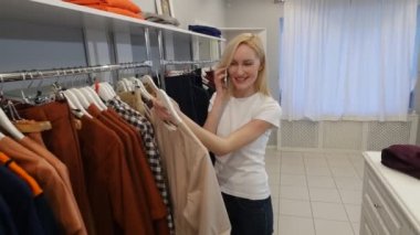 Woman Chooses a Beige Dress Talking by Phone Womenswear Shop Client Blonde Woman is Buying a Clothes in a Boutique Garments Are Hanging on a Trempels