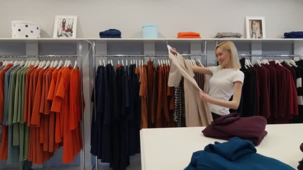 Mujer eligió un vestido beige entre una ropa sonriente va a probarlo Womenswear Shop Cliente Rubia Mujer está comprando una ropa Las prendas están en un temblor — Vídeos de Stock