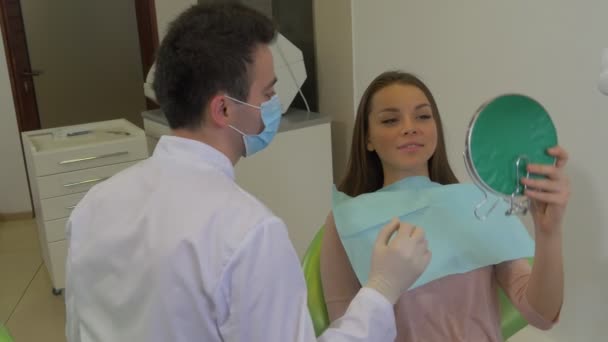 Dentista está probando un nuevo color de dientes para el cliente Mujer sonriente está mirando el espejo sentado en la silla del dentista Estomatólogo masculino en la clínica dental Lab Coat — Vídeos de Stock