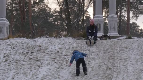 Mamá e hijo en una colina en el parque Bucha Ucrania Navidad sonriendo a su hijo escalando cuesta arriba por nieve niño diapositivas caídas abajo invierno padres están en una colina — Vídeo de stock