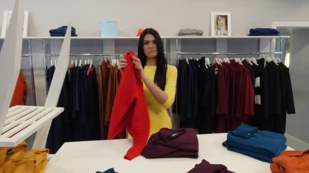 Mujer intenta un suéter rojo mirando el espejo sonriendo cliente morena mujer está comprando una ropa en una ropa de mujer Boutique Garments Are on a Hangers — Vídeos de Stock