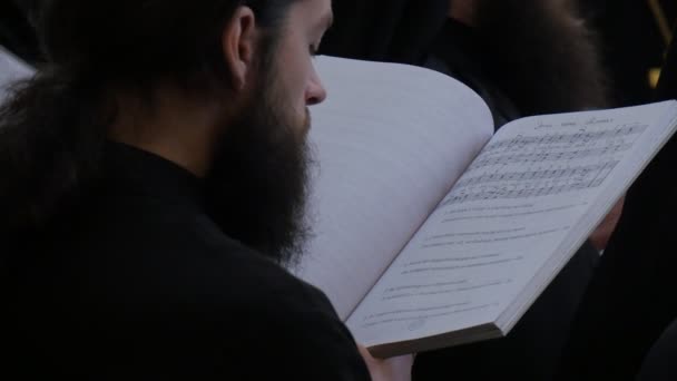 聖職者は、ひげを持つソングブック若い聖職者を保持する黒い衣服でクリスマス聖山ラブラドルミネーション大聖堂ウクライナの男性を歌っています — ストック動画