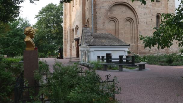 聖職者は、黒いローブの修道士の赤、男性が歩いているコートヤード修道院 Vydubychi 男性修道院で像記念碑近くに歩いて赤れんが造りの教会を残しています。 — ストック動画