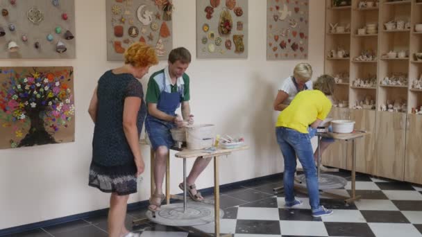 Les gens à la poterie Atelier Master Class Travailler sur une roue de potier Atelier enseignant en jaune aide la femme à faire pivoter sa roue par jambe Femme vient aider — Video