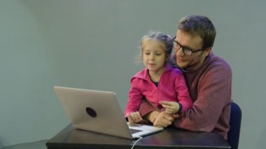 Çocuk Laptop baba Hugs, dizlerinin üzerinde konuşurken bir kızı tutan adam kızı Laptop adam ile çalışma öğretir ve çocuk masada oturuyor