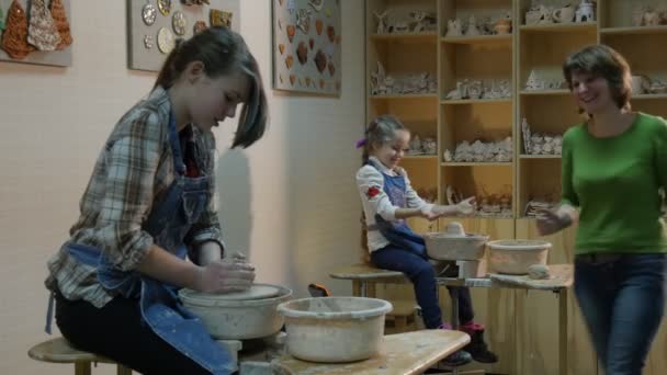 Due ragazze dei bambini sono alle ruote di ceramica insegnante li aiuta a ruotare una ruota con la gamba a parlare con le ragazze Gli studenti sorridenti stanno imparando con entusiasmo — Video Stock
