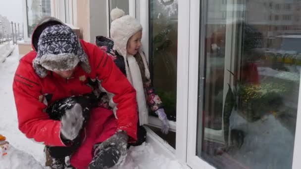 Pai sacode uma neve fora das luvas de uma criança botas sorridente pai e sua filha jogar com neve na janela perto de uma casa em uma cornija ao ar livre inverno nevado — Vídeo de Stock