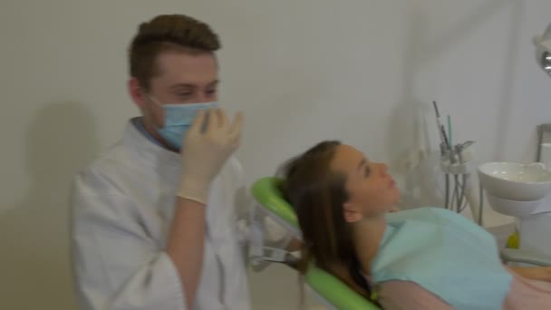 Il dentista sta parlando con una cliente donna sta sorridendo seduto su una sedia verde medico in una maschera cappotto di laboratorio e guanti esaminando una stanza di trattamento dentale dei denti — Video Stock
