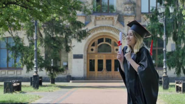 Junge Frau umarmt die glückliche Absolventin und Studentin im Mantel geht vom alten Backsteingebäude der Universität weg, umgeben von parkgrünen Bäumen — Stockvideo