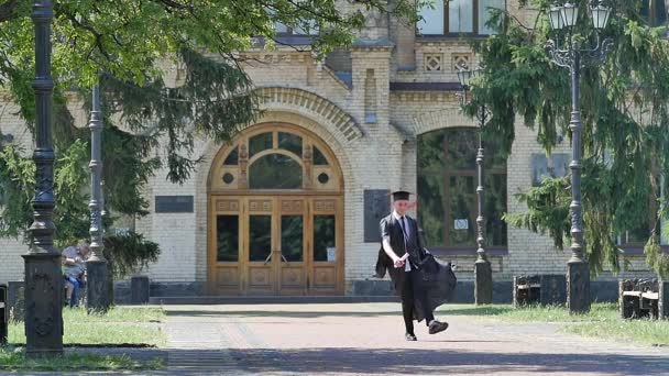 Heureux diplômé sauts jette chapeau marche Université Cour Jeune homme dans le manteau étudiant vieux bâtiment de brique entouré de parc ensoleillé arbres verts — Video