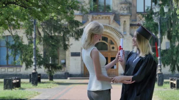 Jeune femme félicite le diplômé dans le manteau debout à l'université deux femmes parlent étreinte bâtiment entouré de parc ensoleillé vert arbres — Video