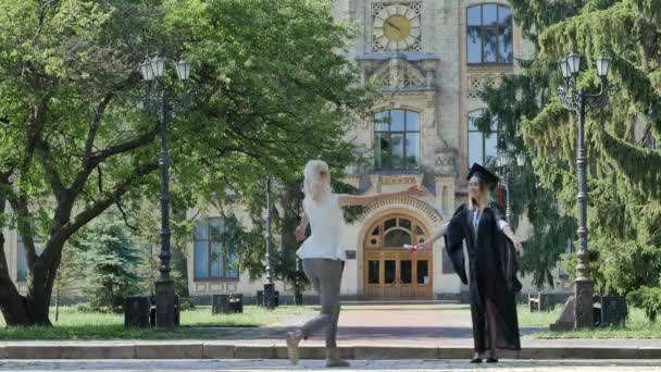 Graduado se encuentra con el amigo y abrazar a pie lejos del manto de la Universidad y el sombrero viejo edificio de ladrillo rodeado de árboles verdes de día soleado Parque — Vídeo de stock