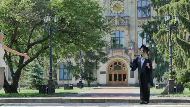 Młoda kobieta jest Hugging uśmiechający się Graduand Student w płaszcz jest chodzenie daleko od Uniwersytetu cegły budynek otoczony z Park zielony starodrzewiem — Wideo stockowe