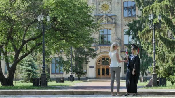 Glada Student i mantel är kramas ung kvinna slutförandet av utbildning gå bort från universitet byggnad omgiven med soliga dag Green parkträd — Stockvideo