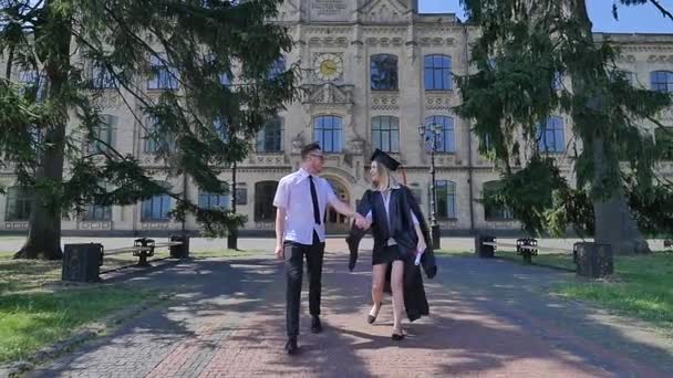 Jeune femme diplômée à Manteau avec jeune homme marchant ensemble tenant la main Vieil immeuble en brique de l'université entouré d'arbres verts Park Sunny Day — Video