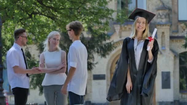 Mujer graduada en manto con diploma sonriente muestra su pulgar parado en el callejón fuera de la universidad Feliz día de graduados detrás de la mujer soleada — Vídeos de Stock