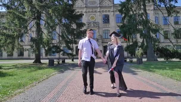 Graduands que tem o homem novo do divertimento que torce uma mulher no envoltório estudantes felizes que sorriem andando pela aléia fora da terra arrendada da Universidade entrega o dia de verão ensolarado — Vídeo de Stock