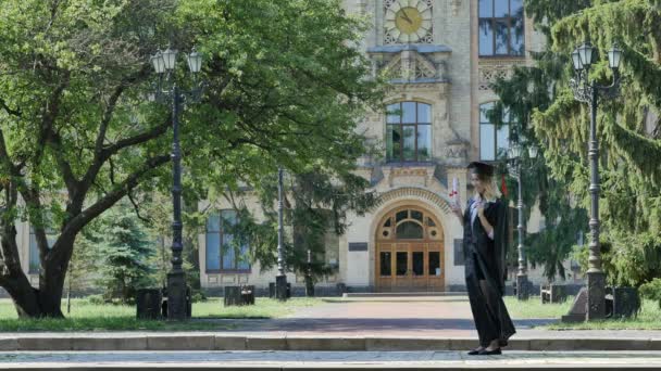 Femme félicite Graduand étreint fille dans le manteau étudiant montre son diplôme souriant secoue le papier debout dans l'allée à l'extérieur de l'université Journée ensoleillée — Video