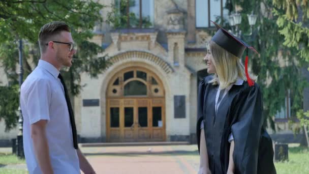 Femme Diplômée et souriante Coquettishly montre Diplôme Homme vient sourire Embrasse la femme debout dans l'allée devant l'université Heureux diplômés dans la journée ensoleillée — Video