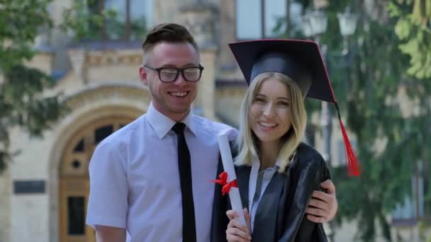 Пара щасливих жінок випускників показує її диплом усміхнений чоловік в сорочці і краватку обіймає жінку в мантії стоячи на алеї за межами університету в сонячний день — стокове відео