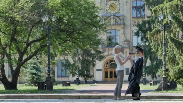 Woman Comgretulates Graduand Girl in Mantle Hugs Her Girl Holding Diploma Smiling Talking to Friend Mom Sister Alley Outside the University Sunny Day — ストック動画