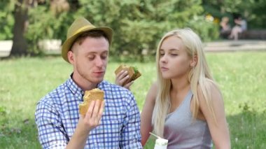 Young Couple Eating Sandwiches on a Lawn Talking Smiling Drinking Juice Picnic People Sitting on a Green Grass in Park Happy People Sunny Summer Day