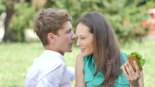 Couple Girl is Eating Sandwich on a Lawn Talking Smiling Couple at the Picnic Man and Woman Sitting on a Green Grass in Park Happy People Sunny Summer Day — Stock Video