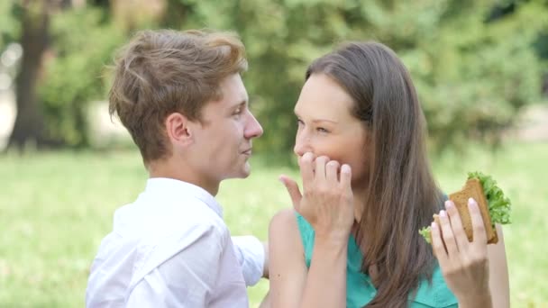 Joven mujer está comiendo sándwich en un césped hablando sonriente pareja en el picnic feliz hombre y mujer sentado en una hierba verde en el parque soleado día de verano — Vídeos de Stock