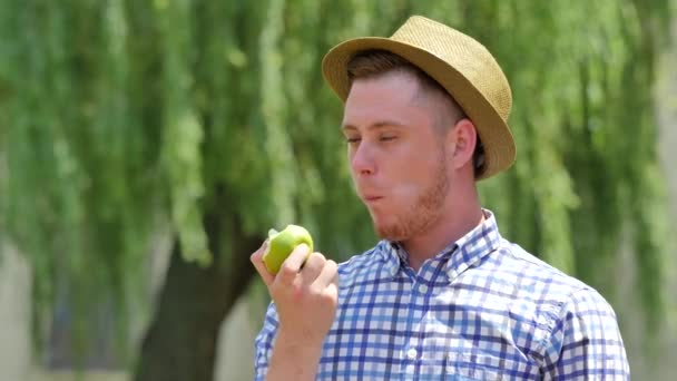 Young Man Smells and Eating an Apple Smiling Outdoors Willow Tree Branches Are Swaying at the Wind Guy in Checkered Shirt and Hat Bites Green Apple Sunny — Wideo stockowe