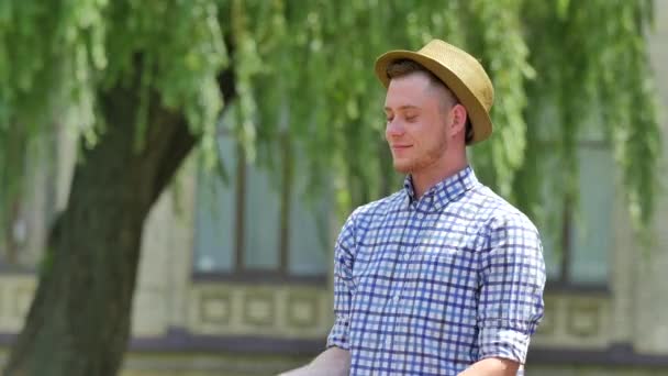 Young Man Throws an Apple up Smells Eats Smiling Outdoors Willow Tree Branches Are Swaying at the Wind Guy in Checkered Shirt and Hat Bites Green Apple — Stock video