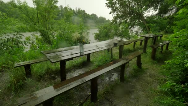 Antiguas mesas y bancos de madera en la lluvia vertida Base turística Árboles verdes exuberantes Lluvia forestal está lavando todo rastro Día nublado lluvioso de verano en el río — Vídeo de stock