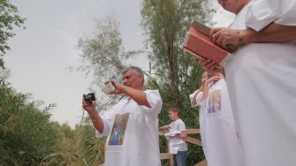 Män som tar bilder av kvinnor under dopet Rite Jordan River Israel liten sumpig flod Clayish botten trätrappor till vattnet grönt gräs vass träd — Stockvideo