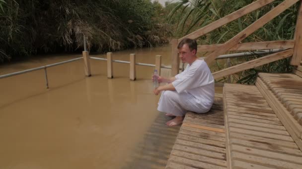 Muž sedící na schodech v jordánském břehu Jordánu baptismus muž bere vodu do láhve popíjet malou Svastovou řeku dřevěné schody k vodě — Stock video