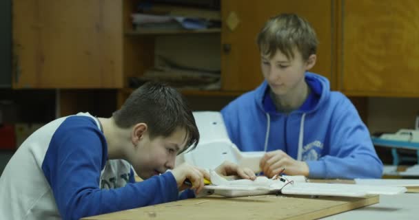 Δύο εφηβικά αγόρια κάνουν λεπτομέρειες για το αεροπλάνο, κρατώντας το φτιαγμένο αεροπλάνο, χαμογελώντας, καθιστός στο τραπέζι, παιδικό εργαστήρι μοντελοποίησης παιδιών — Αρχείο Βίντεο
