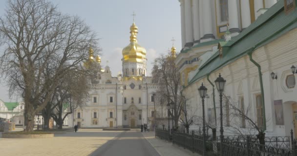 Люди йдуть поруч з Успенським собором Києво-Печерської Лаври золотий купол без листя білі стіни вуличні світильники блакитне небо сонячно — стокове відео