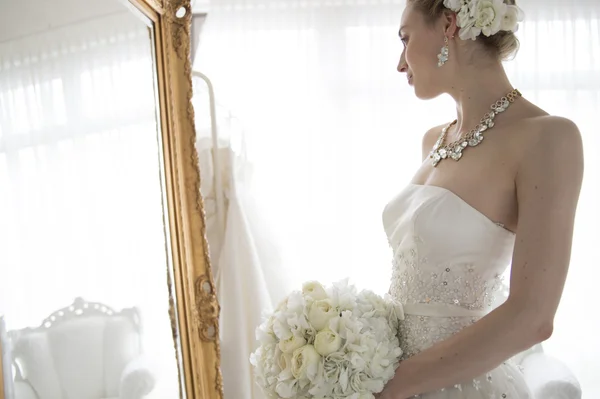 Menina bonita noiva morena em vestido de noiva branco com penteado e maquiagem brilhante olha no espelho — Fotografia de Stock