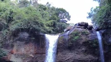 Karanlık kayalık uçurumlarda çağlayan tropikal bir şelalenin sakin 4K görüntüsü birden fazla akarsu ve parlak doğal gökyüzü altında canlı yeşil yapraklar barındıran bereketli orman havzasına dönüşüyor.