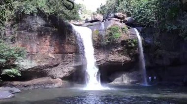 Sersemletici tropikal şelale kayalık bir uçurumdan serin bir havuza dökülüyor. Yeşillik ve mağara benzeri kaya oluşumu huzurlu ve doğal bir manzara yaratır.