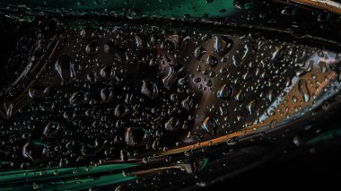 Pürüzsüz, koyu metalik bir yüzey üzerinde su damlacıklarının bu yakın çekim görüntüsü karamsar yeşil ve kehribar yansımaları yüksek kontrastlı, soyut