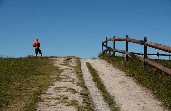 Tepenin tepesinde duran kırmızı kapüşonlu adam. Kumlu yol ünlü Norveç plajı Farstadstranda 'ya çıkar. Mavi gökyüzü arka planı. Boşluğu kopyala.