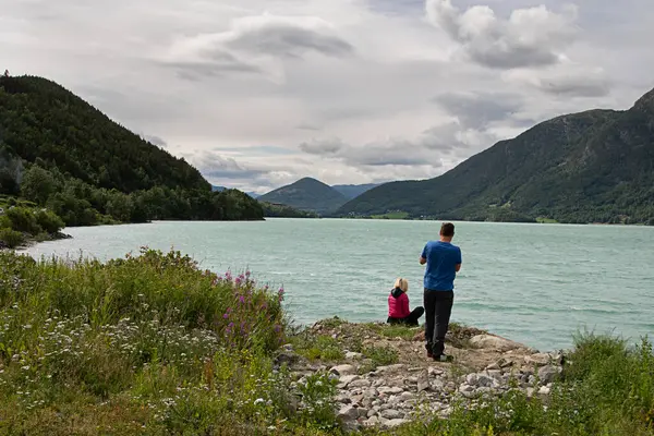 Vgvatnet (lake), Norway- July 21, 2020: Man and woman view the turquoise lake and take photographs with their mobile phones. Mountains in the background. Cloudy sky.