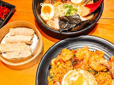 A hearty bowl of delicious fried seafood ramen noodles and beef ramen in tori paitan soup with a side dish of shrimp dim sum on a bamboo tray. Asian cuisine at a restaurant